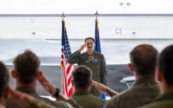 A military officer in a flight suit stands at the front of a hangar, returning a salute to a formation of assembled personnel. The ceremony appears to be a formal military event such as a change of command or unit activation, taking place in an aircraft hangar with a fighter jet visible in the background. Both the American flag and what appears to be a U.S. Air Force flag are prominently displayed behind the officer.