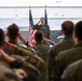 A military officer in a flight suit stands at the front of a hangar, returning a salute to a formation of assembled personnel. The ceremony appears to be a formal military event such as a change of command or unit activation, taking place in an aircraft hangar with a fighter jet visible in the background. Both the American flag and what appears to be a U.S. Air Force flag are prominently displayed behind the officer.