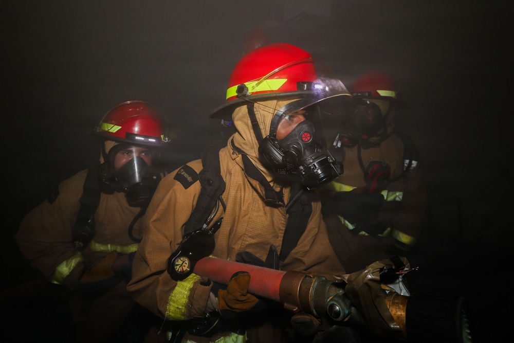 Three firefighters are operating in a smoke-filled environment, wearing full personal protective equipment including turnout gear and respiratory protection. The lead firefighter is gripping a fire hose nozzle while advancing through heavy smoke. The scene appears to be an active firefighting operation or training exercise conducted in low-visibility conditions.