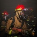 Three firefighters are operating in a smoke-filled environment, wearing full personal protective equipment including turnout gear and respiratory protection. The lead firefighter is gripping a fire hose nozzle while advancing through heavy smoke. The scene appears to be an active firefighting operation or training exercise conducted in low-visibility conditions.