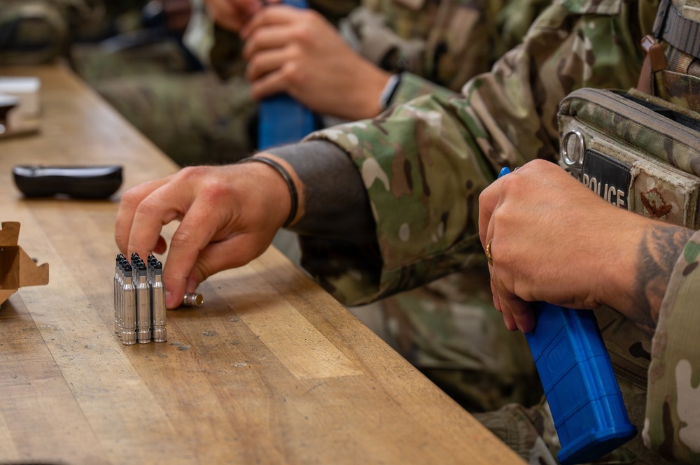 Military or law enforcement personnel in camouflage uniforms are at a wooden table handling ammunition and a blue magazine. A patch reading 'POLICE' is visible on one individual's tactical vest. The scene appears to be a range preparation or weapons qualification activity.