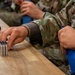 Military or law enforcement personnel in camouflage uniforms are at a wooden table handling ammunition and a blue magazine. A patch reading 'POLICE' is visible on one individual's tactical vest. The scene appears to be a range preparation or weapons qualification activity.