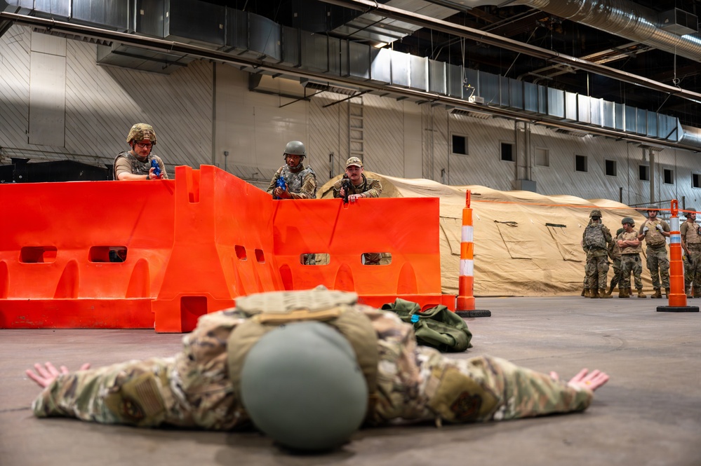 Military personnel are conducting an indoor training exercise in a large warehouse or hangar facility. One soldier is prone on the ground with hands spread flat, while two others take cover positions behind orange plastic barriers aiming training weapons. Additional personnel observe the exercise in the background near tan military tents.