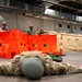 Military personnel are conducting an indoor training exercise in a large warehouse or hangar facility. One soldier is prone on the ground with hands spread flat, while two others take cover positions behind orange plastic barriers aiming training weapons. Additional personnel observe the exercise in the background near tan military tents.