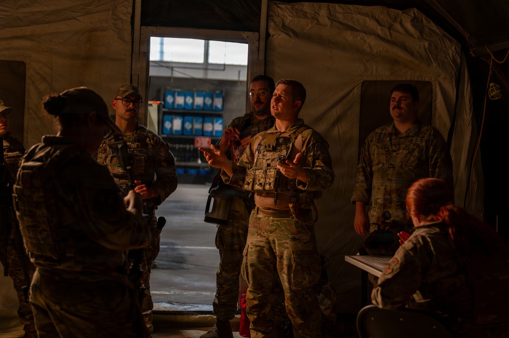 A group of military personnel in camouflage uniforms are gathered inside what appears to be a military tent or temporary structure. One individual in the center, wearing a tactical vest, appears to be briefing or instructing the group with hand gestures. The scene is dimly lit with an open doorway providing background light revealing an outdoor area.