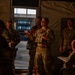 A group of military personnel in camouflage uniforms are gathered inside what appears to be a military tent or temporary structure. One individual in the center, wearing a tactical vest, appears to be briefing or instructing the group with hand gestures. The scene is dimly lit with an open doorway providing background light revealing an outdoor area.