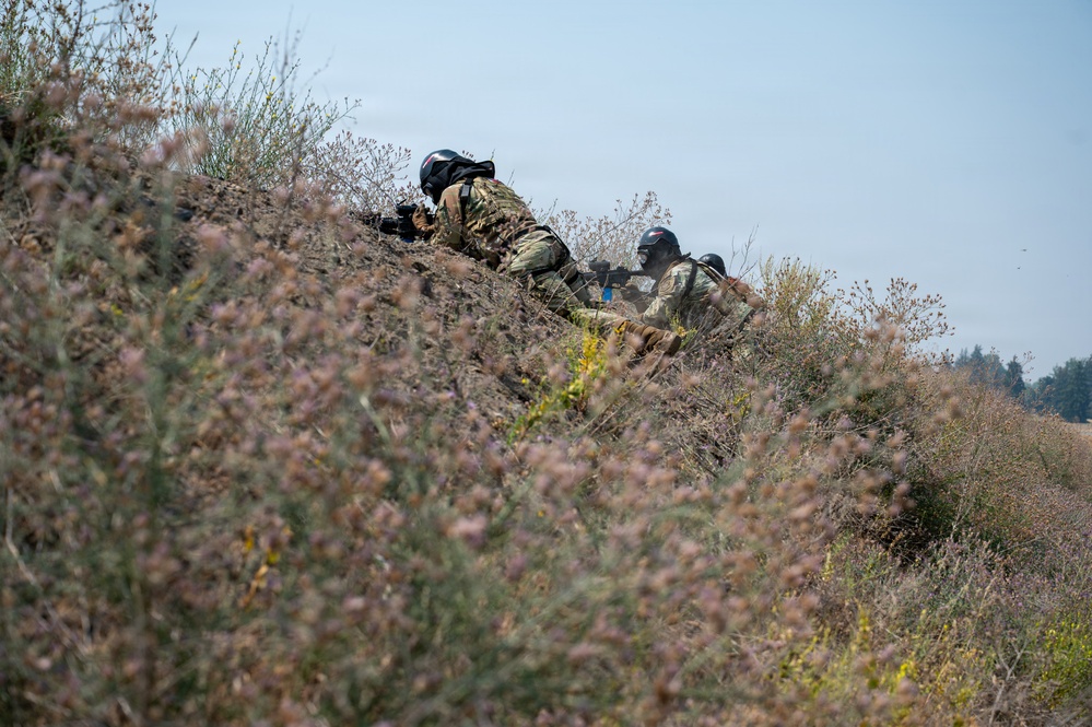 Two individuals in military-style camouflage uniforms and black helmets are positioned prone on a dirt embankment or berm, aiming rifles in a forward direction. They are partially concealed by wild vegetation with small pink/purple flowers in the foreground. The scene appears to be an outdoor tactical exercise or field operation.