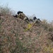 Two individuals in military-style camouflage uniforms and black helmets are positioned prone on a dirt embankment or berm, aiming rifles in a forward direction. They are partially concealed by wild vegetation with small pink/purple flowers in the foreground. The scene appears to be an outdoor tactical exercise or field operation.