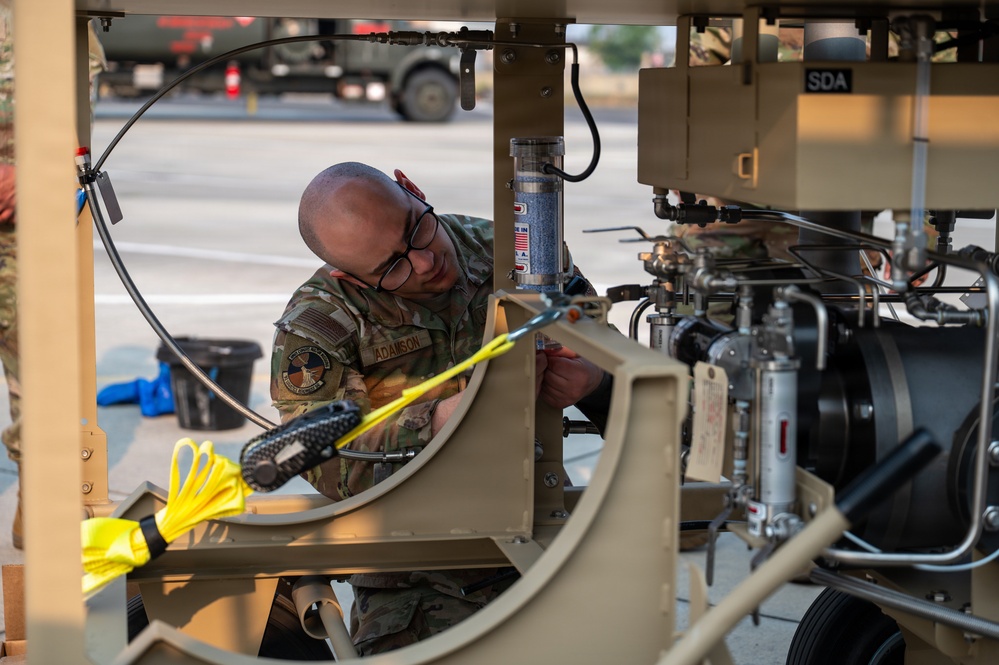 A military service member identified by name tape as 'ADAMSON' is performing maintenance or inspection work on a piece of military ground support equipment. The individual is wearing OCP (Operational Camouflage Pattern) uniform and glasses, working within the frame structure of what appears to be a tan-colored military support vehicle or equipment unit. The setting appears to be an outdoor military installation or flight line area.