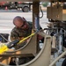 A military service member identified by name tape as 'ADAMSON' is performing maintenance or inspection work on a piece of military ground support equipment. The individual is wearing OCP (Operational Camouflage Pattern) uniform and glasses, working within the frame structure of what appears to be a tan-colored military support vehicle or equipment unit. The setting appears to be an outdoor military installation or flight line area.