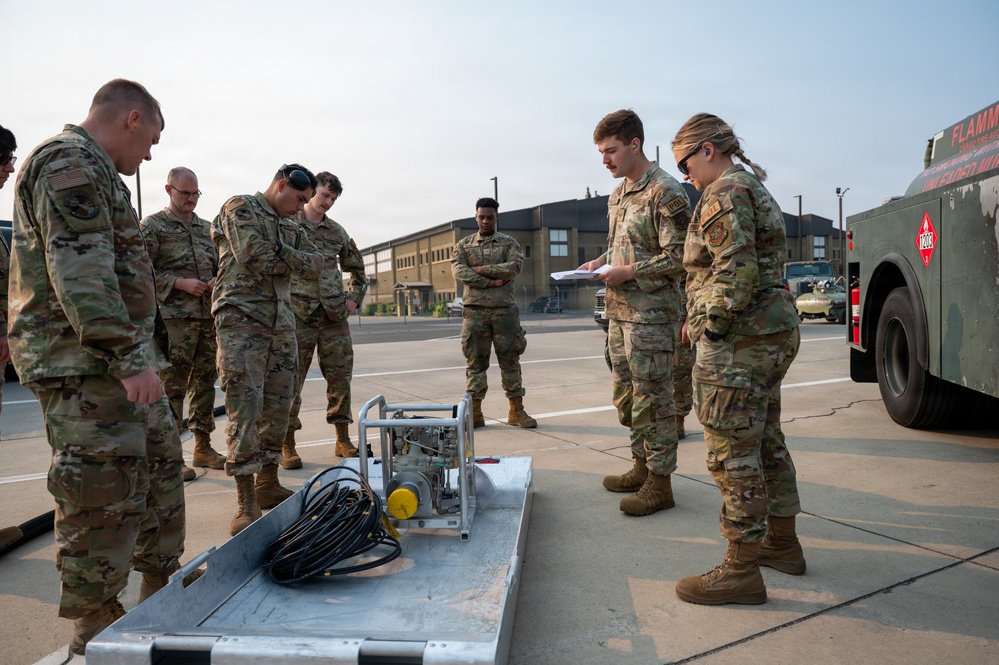 A group of military personnel in OCP (Operational Camouflage Pattern) uniforms are gathered outdoors on a paved surface, appearing to conduct a training or briefing session. They are focused on a piece of equipment laid out on a metal platform, which includes cables and mechanical components. A fuel/flammable materials truck is visible in the background to the right.