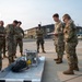 A group of military personnel in OCP (Operational Camouflage Pattern) uniforms are gathered outdoors on a paved surface, appearing to conduct a training or briefing session. They are focused on a piece of equipment laid out on a metal platform, which includes cables and mechanical components. A fuel/flammable materials truck is visible in the background to the right.