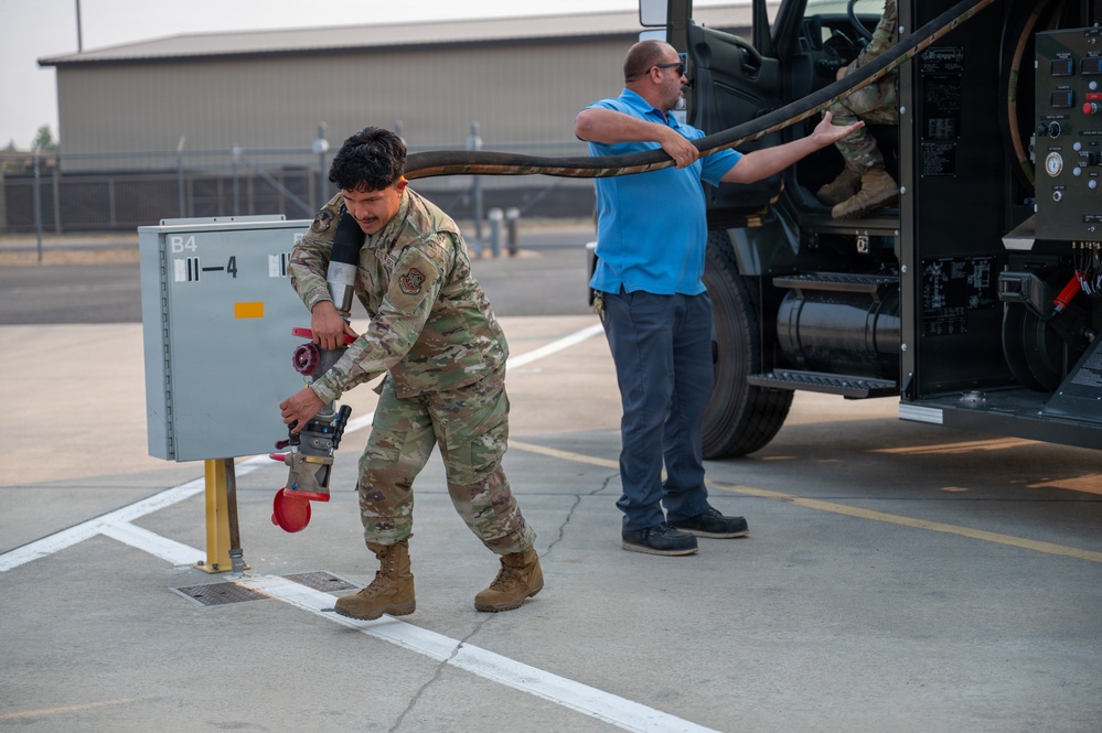 Two personnel are working on a military airfield apron, with one individual in camouflage uniform connecting or disconnecting a large hose to a ground utility panel labeled 'B4' and '11-4'. A second person in a blue civilian shirt assists by managing the large hose extending from a military support vehicle. The setting appears to be a flight line at a U.S. Air Force installation.