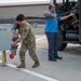 Two personnel are working on a military airfield apron, with one individual in camouflage uniform connecting or disconnecting a large hose to a ground utility panel labeled 'B4' and '11-4'. A second person in a blue civilian shirt assists by managing the large hose extending from a military support vehicle. The setting appears to be a flight line at a U.S. Air Force installation.