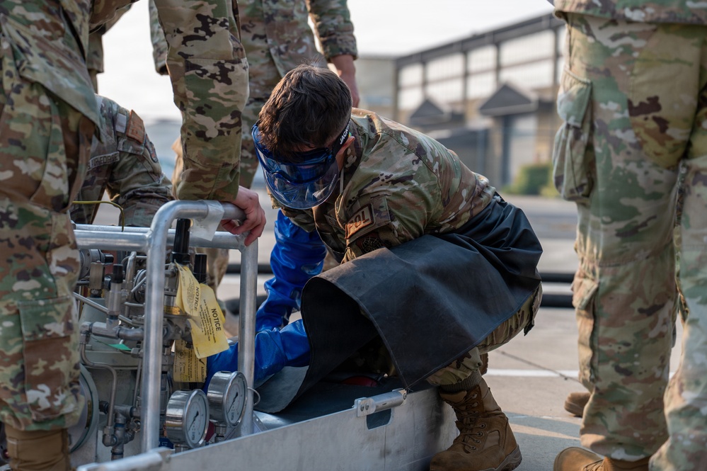 Military personnel in OCP (Operational Camouflage Pattern) uniforms are gathered around a piece of equipment, likely during a training exercise. One service member wearing a face shield and protective apron is crouched down, closely inspecting or operating a pressurized equipment cart with gauges and yellow warning tags. The setting appears to be an outdoor military installation or airfield.