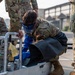 Military personnel in OCP (Operational Camouflage Pattern) uniforms are gathered around a piece of equipment, likely during a training exercise. One service member wearing a face shield and protective apron is crouched down, closely inspecting or operating a pressurized equipment cart with gauges and yellow warning tags. The setting appears to be an outdoor military installation or airfield.