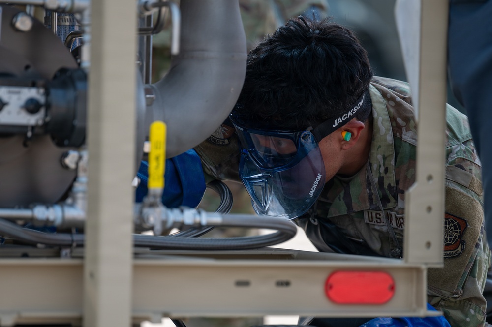 A U.S. Air Force service member identified as 'Jackson' is closely inspecting or performing maintenance on a piece of military equipment. The individual is wearing full personal protective equipment including blue safety goggles, blue gloves, and orange ear protection. The equipment appears to be a fuel or fluid handling system mounted on a trailer frame.