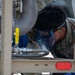 A U.S. Air Force service member identified as 'Jackson' is closely inspecting or performing maintenance on a piece of military equipment. The individual is wearing full personal protective equipment including blue safety goggles, blue gloves, and orange ear protection. The equipment appears to be a fuel or fluid handling system mounted on a trailer frame.