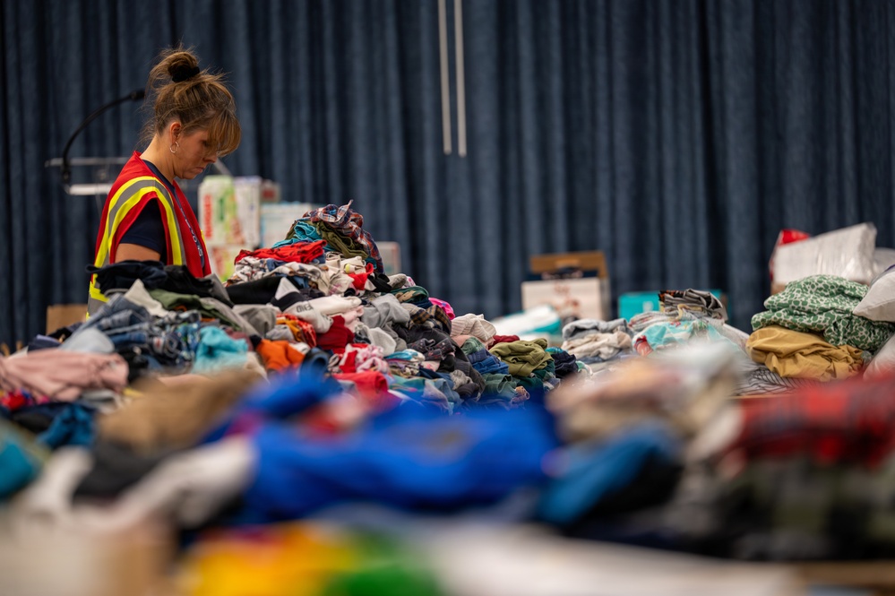 A volunteer or worker wearing a high-visibility safety vest is sorting through large piles of donated clothing inside what appears to be a community center or gymnasium. The scene suggests a disaster relief or charity donation operation, with massive quantities of mixed clothing spread across tables. The blue curtain backdrop and indoor setting indicate an organized collection and distribution effort.