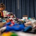 A volunteer or worker wearing a high-visibility safety vest is sorting through large piles of donated clothing inside what appears to be a community center or gymnasium. The scene suggests a disaster relief or charity donation operation, with massive quantities of mixed clothing spread across tables. The blue curtain backdrop and indoor setting indicate an organized collection and distribution effort.