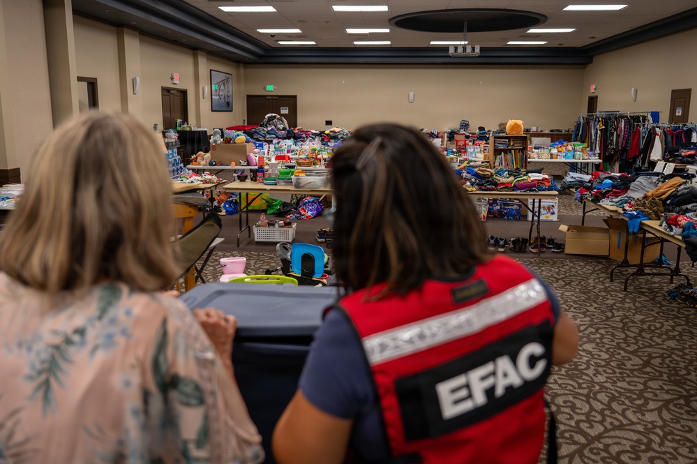 Two women are standing at the entrance of a large indoor relief or donation distribution center. One woman is wearing a red vest labeled 'EFAC', indicating an emergency or disaster relief organization. The room is filled with tables covered in donated goods including clothing, toys, shoes, and household items.