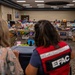 Two women are standing at the entrance of a large indoor relief or donation distribution center. One woman is wearing a red vest labeled 'EFAC', indicating an emergency or disaster relief organization. The room is filled with tables covered in donated goods including clothing, toys, shoes, and household items.