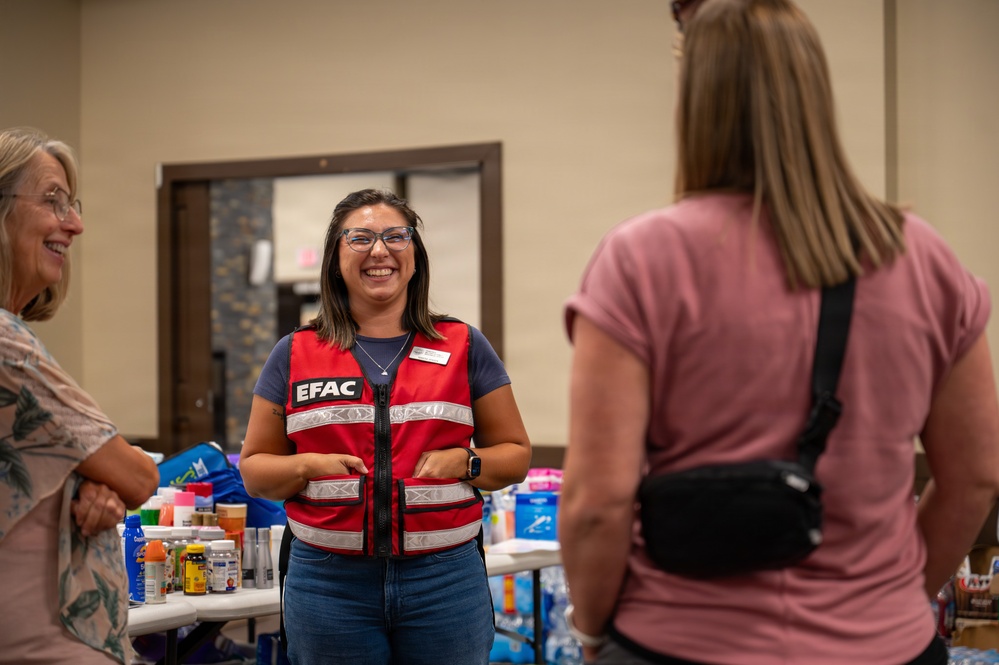 A smiling woman wearing a red 'EFAC' vest is engaged in conversation with two other women at what appears to be a community relief or emergency assistance distribution event. Tables behind her are stocked with various supplies including medications, water bottles, and other essential items. The setting appears to be an indoor community hall or church facility.