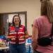 A smiling woman wearing a red 'EFAC' vest is engaged in conversation with two other women at what appears to be a community relief or emergency assistance distribution event. Tables behind her are stocked with various supplies including medications, water bottles, and other essential items. The setting appears to be an indoor community hall or church facility.