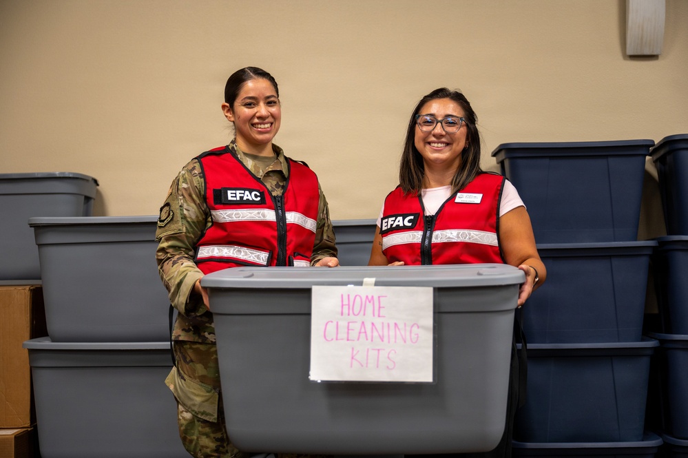 Two women wearing red 'EFAC' vests are holding a gray plastic storage bin labeled 'Home Cleaning Kits' and smiling at the camera. One woman is dressed in military camouflage uniform while the other wears civilian clothing. They appear to be participating in a disaster relief or community assistance effort, surrounded by multiple storage bins.