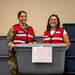 Two women wearing red 'EFAC' vests are holding a gray plastic storage bin labeled 'Home Cleaning Kits' and smiling at the camera. One woman is dressed in military camouflage uniform while the other wears civilian clothing. They appear to be participating in a disaster relief or community assistance effort, surrounded by multiple storage bins.