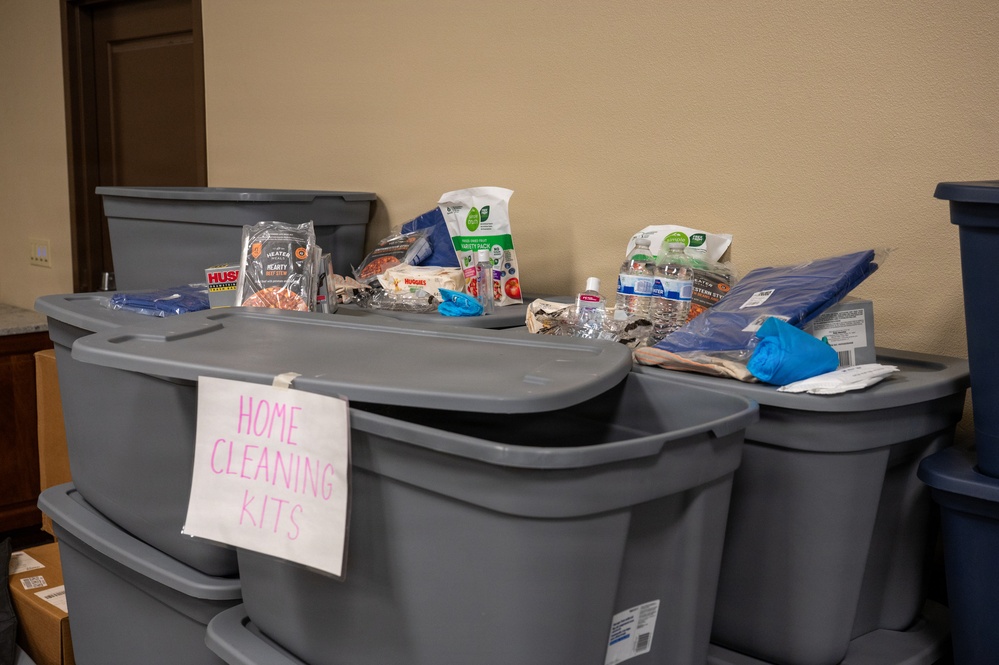 The image shows a collection of large gray plastic storage bins labeled 'HOME CLEANING KITS' in pink handwritten text. The bins are stacked and filled with various household cleaning and hygiene supplies, suggesting a community aid or disaster relief distribution effort. Multiple items are visible on top of and inside the open bins.