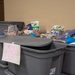 The image shows a collection of large gray plastic storage bins labeled 'HOME CLEANING KITS' in pink handwritten text. The bins are stacked and filled with various household cleaning and hygiene supplies, suggesting a community aid or disaster relief distribution effort. Multiple items are visible on top of and inside the open bins.