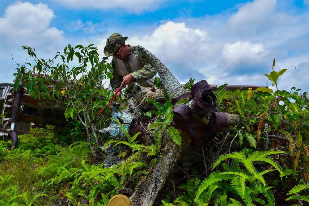 A person wearing camouflage clothing and a cap is examining heavily corroded and overgrown metal wreckage, likely remnants of military equipment or aircraft. The wreckage is extensively covered in rust, patina, and tropical vegetation including ferns and small trees. The setting appears to be a tropical or subtropical environment with lush greenery and a partly cloudy sky.