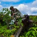 A person wearing camouflage clothing and a cap is examining heavily corroded and overgrown metal wreckage, likely remnants of military equipment or aircraft. The wreckage is extensively covered in rust, patina, and tropical vegetation including ferns and small trees. The setting appears to be a tropical or subtropical environment with lush greenery and a partly cloudy sky.