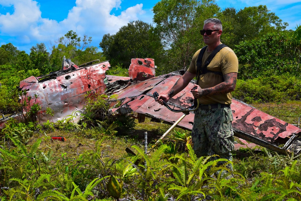 A military personnel member in camouflage pants and olive t-shirt stands before a heavily deteriorated aircraft wreck overgrown with tropical vegetation. The wreckage, painted red and riddled with holes and corrosion damage, appears to be a World War II-era aircraft in an advanced state of decay. The setting suggests a Pacific island location, consistent with WWII battlefield archaeology or recovery operations.