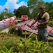 A military personnel member in camouflage pants and olive t-shirt stands before a heavily deteriorated aircraft wreck overgrown with tropical vegetation. The wreckage, painted red and riddled with holes and corrosion damage, appears to be a World War II-era aircraft in an advanced state of decay. The setting suggests a Pacific island location, consistent with WWII battlefield archaeology or recovery operations.