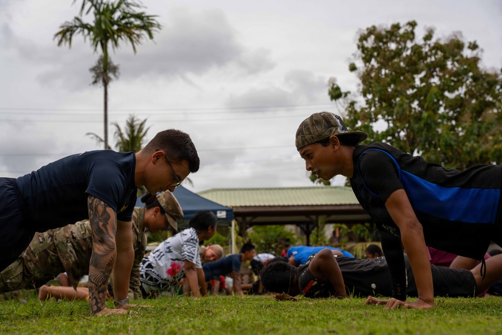 A group of individuals are performing push-ups on a grass field in an outdoor setting. The foreground features two men facing each other in the push-up position, one wearing a navy blue shirt with visible tattoos and glasses, and another wearing a black and blue shirt with a camouflage cap. In the background, additional participants and what appears to be a military or uniformed individual in camouflage are also doing push-ups near a covered pavilion structure.
