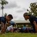 A group of individuals are performing push-ups on a grass field in an outdoor setting. The foreground features two men facing each other in the push-up position, one wearing a navy blue shirt with visible tattoos and glasses, and another wearing a black and blue shirt with a camouflage cap. In the background, additional participants and what appears to be a military or uniformed individual in camouflage are also doing push-ups near a covered pavilion structure.