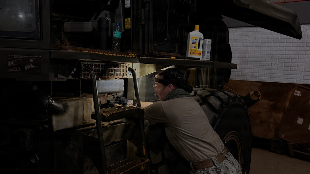 A person wearing military-style camouflage pants and a tan shirt is performing maintenance or inspection work on a large military vehicle, likely a HMMWV (Humvee) or similar tactical vehicle. The work is taking place in an indoor garage or maintenance bay with a white brick wall visible in the background. The vehicle bears a 'US Department of the Navy' data plate, indicating it is U.S. Navy property.