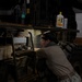 A person wearing military-style camouflage pants and a tan shirt is performing maintenance or inspection work on a large military vehicle, likely a HMMWV (Humvee) or similar tactical vehicle. The work is taking place in an indoor garage or maintenance bay with a white brick wall visible in the background. The vehicle bears a 'US Department of the Navy' data plate, indicating it is U.S. Navy property.