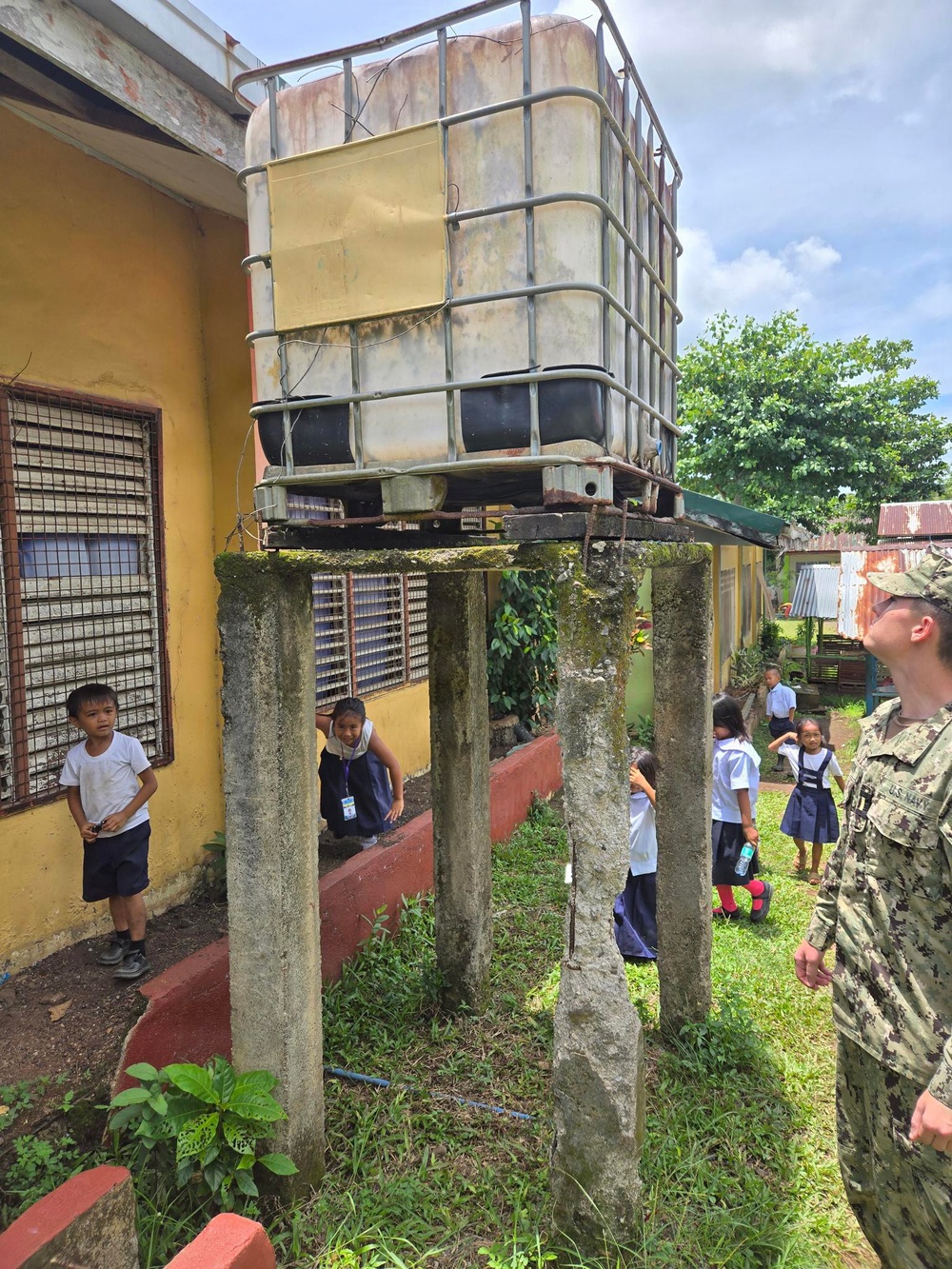 A U.S. Navy service member is inspecting an elevated water storage tank (IBC tote) mounted on deteriorating concrete pillars at what appears to be a school compound. Several children in school uniforms are present in the area, suggesting this may be a humanitarian assistance or civic action visit. The infrastructure shows significant weathering and aging, with moss-covered concrete supports and a rusted metal platform.