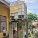 A U.S. Navy service member is inspecting an elevated water storage tank (IBC tote) mounted on deteriorating concrete pillars at what appears to be a school compound. Several children in school uniforms are present in the area, suggesting this may be a humanitarian assistance or civic action visit. The infrastructure shows significant weathering and aging, with moss-covered concrete supports and a rusted metal platform.
