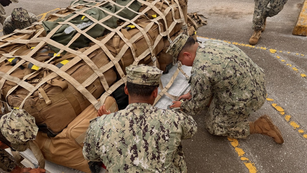 Several military personnel in digital camouflage uniforms are working together to secure or inspect a large cargo bundle wrapped in a tan cargo net on a paved surface. The bundle contains multiple tactical backpacks and gear bags of various colors. The scene appears to take place on a flight deck, tarmac, or staging area marked with yellow lines.