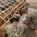 Several military personnel in digital camouflage uniforms are working together to secure or inspect a large cargo bundle wrapped in a tan cargo net on a paved surface. The bundle contains multiple tactical backpacks and gear bags of various colors. The scene appears to take place on a flight deck, tarmac, or staging area marked with yellow lines.