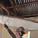 A military personnel member wearing U.S. Navy digital camouflage (NWU Type III) and a hard hat is working inside a severely damaged building, appearing to handle electrical wiring in the ceiling area. The structure shows significant damage with an exposed corrugated metal roof, deteriorated ceiling, and damaged walls. This appears to be a disaster relief or humanitarian assistance operation.