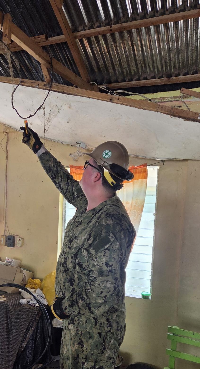 A military personnel member wearing U.S. Navy digital camouflage (NWU Type III) and a hard hat is working inside a severely damaged building, appearing to handle electrical wiring in the ceiling area. The structure shows significant damage with an exposed corrugated metal roof, deteriorated ceiling, and damaged walls. This appears to be a disaster relief or humanitarian assistance operation.