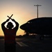 A ground crew member (marshaller) is silhouetted against a sunset sky on an airport tarmac, signaling a large aircraft with crossed wands raised above their head. The dramatic backlighting creates a strong silhouette effect with warm orange and yellow tones from the setting or rising sun. The scene appears to be at a military or commercial airfield during dusk or dawn operations.