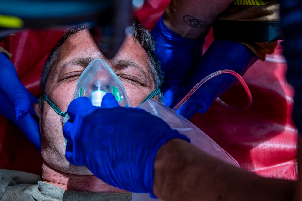 A middle-aged male patient is receiving emergency medical treatment, with an oxygen mask being applied to his face by at least two responders wearing blue nitrile gloves. The patient appears unconscious or semi-conscious with eyes closed, lying on what appears to be a red stretcher or backboard. The scene is consistent with a pre-hospital emergency medical response.