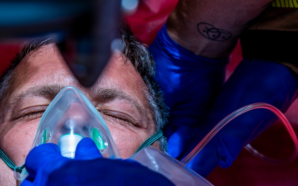 A middle-aged male patient is receiving emergency medical treatment, with an oxygen mask being applied to his face by at least two responders wearing blue nitrile gloves. The patient appears unconscious or semi-conscious with eyes closed, lying on what appears to be a red stretcher or backboard. The scene is consistent with a pre-hospital emergency medical response.