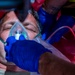 A middle-aged male patient is receiving emergency medical treatment, with an oxygen mask being applied to his face by at least two responders wearing blue nitrile gloves. The patient appears unconscious or semi-conscious with eyes closed, lying on what appears to be a red stretcher or backboard. The scene is consistent with a pre-hospital emergency medical response.